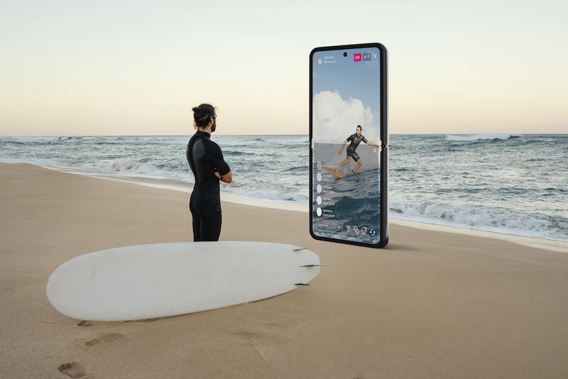 Man som står på strand med med en enorm telefon framför sig med bild på en surfare